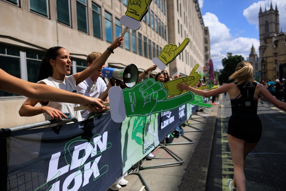 A runner participating in a race is greeted with enthusiasm by spectators along the course. The spectators, standing behind a barrier adorned with a "LDN 10K" banner, cheer with foam hands and a megaphone in hand. The scene is set on a sunny day, with buildings and part of a cathedral visible in the background.