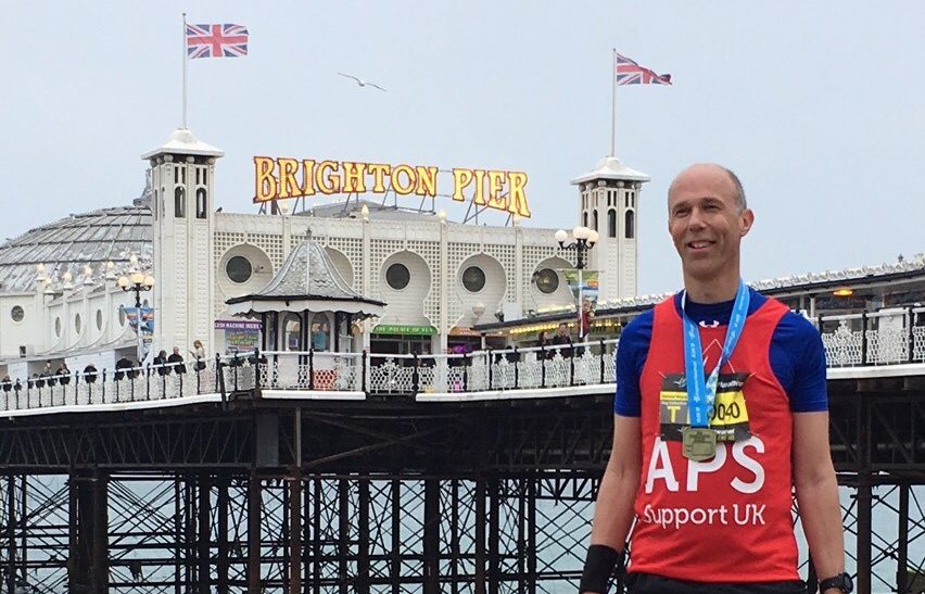 A runner stands proudly on the pebble beach in front of Brighton Pier, wearing a red APS Support UK vest and showcasing their race medals. The iconic pier, with its Union Jack flags and lit-up "Brighton Pier" sign, serves as a striking backdrop. The scene captures a moment of achievement and support for the charity.