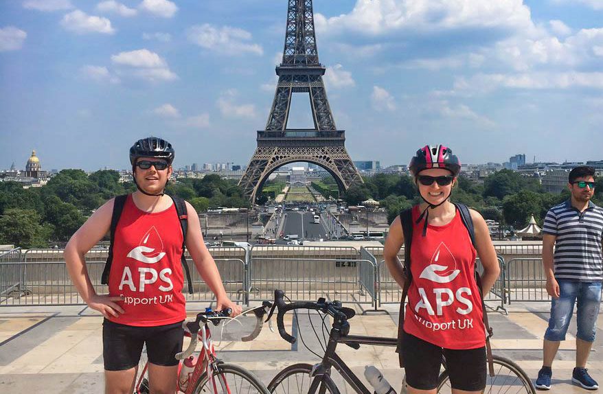Two cyclists stand in front of the Eiffel Tower in Paris, wearing matching red APS Support UK vests. They are posing with their bikes under a clear blue sky, with other people and the cityscape in the background.