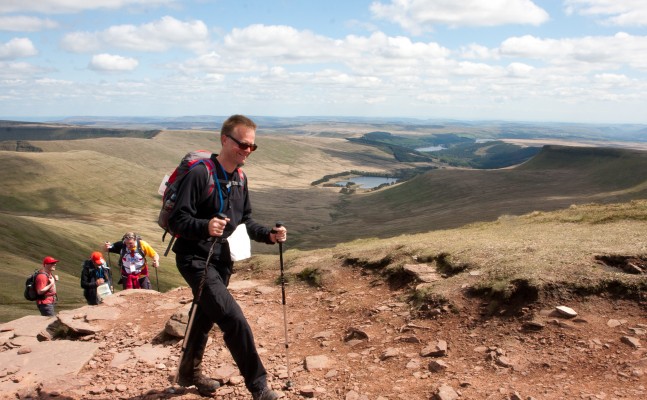 A hiker ascending a rocky mountain trail, using trekking poles, wearing a backpack and sunglasses. Behind them, a group of hikers follows the same path. The landscape features rolling green hills, a distant lake, and expansive views under a bright blue sky dotted with fluffy white clouds.