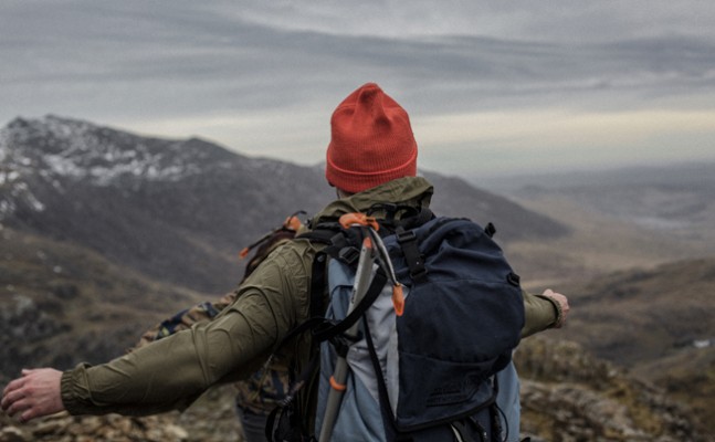 A person standing on a rocky mountain trail with their arms outstretched, wearing a red beanie, a green jacket, and a large hiking backpack. The individual faces away from the camera, overlooking a dramatic landscape of rugged hills and valleys under a cloudy sky, with patches of snow visible on distant peaks.