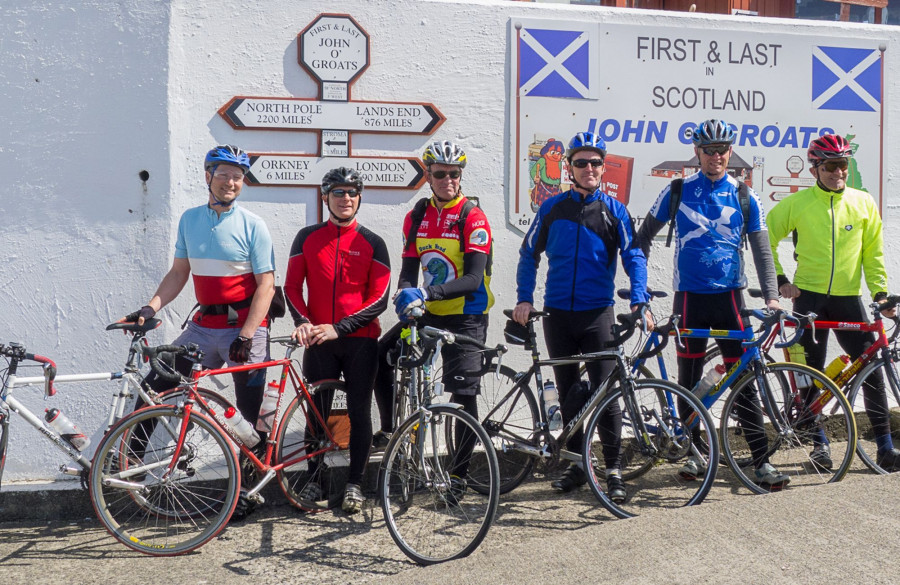 A group of six cyclists standing with their bicycles in front of the iconic signpost at John o' Groats in Scotland. The signpost displays distances to notable locations, including Land’s End (876 miles) and the North Pole (2200 miles). Each cyclist is wearing a colourful cycling outfit and helmet, with a variety of bicycles parked in front of them. Two Scottish flags are visible on either side of the signpost, along with a board reading "First & Last in Scotland John o' Groats."