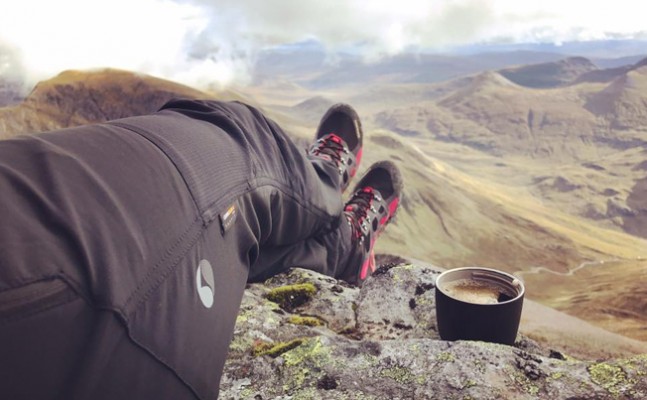 A relaxing view of a hiker's legs stretched out on a rocky surface, wearing black trousers and hiking boots with red accents. A cup of coffee rests beside them, overlooking a stunning mountainous landscape with rolling hills, valleys, and a partially cloudy sky in the background.