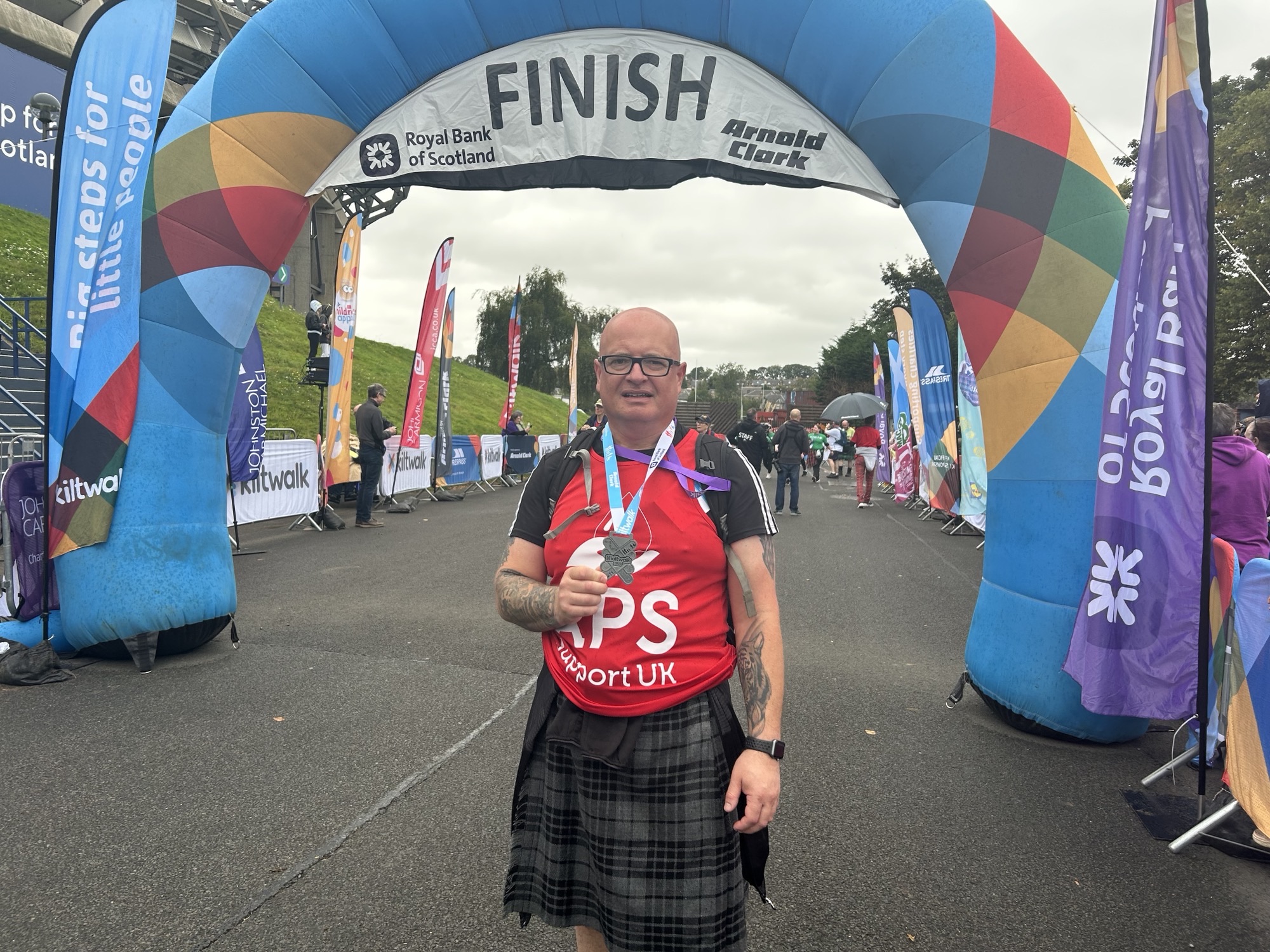 A participant proudly standing under the colourful "Finish" arch of the Kiltwalk event, wearing a red APS Support UK t-shirt and a kilt. They are holding a medal and smiling, with event banners and other participants visible in the background on an overcast day. The scene celebrates their accomplishment and the vibrant atmosphere of the event.