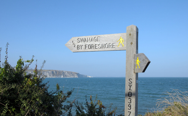 A wooden signpost by the seaside with two directional arrows. One arrow points left and reads "1/2 Swanage by Foreshore" with a yellow walking figure icon. The other arrow points right with a yellow walking figure icon but no text. In the background, the sea stretches out under a clear blue sky, with cliffs visible in the distance. Bushes and grass frame the base of the signpost.