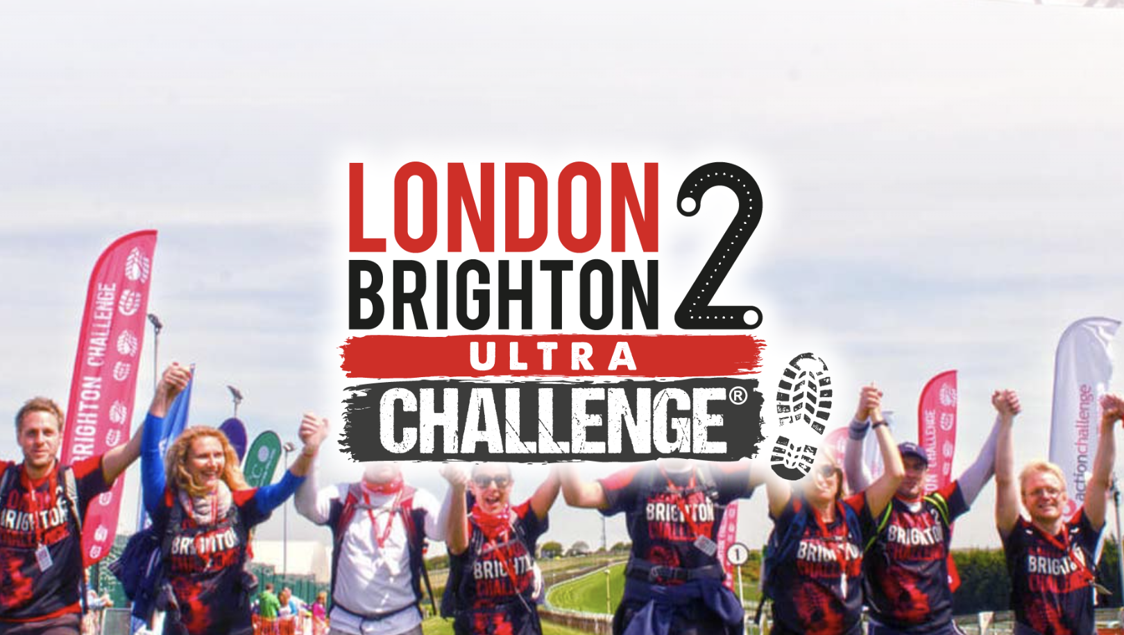 Participants of the London to Brighton Ultra Challenge celebrate crossing the finish line, holding hands in triumph. They wear matching event t-shirts, with vibrant flags and banners waving in the background under a bright sky.