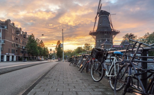 A picturesque street scene at sunset featuring a line of bicycles parked along a railing on a cobblestone sidewalk. To the right, a traditional Dutch windmill stands tall against a dramatic sky filled with golden and orange hues. Trees and buildings line the street on the left, creating a serene urban setting. The warm lighting enhances the charm of the scene.