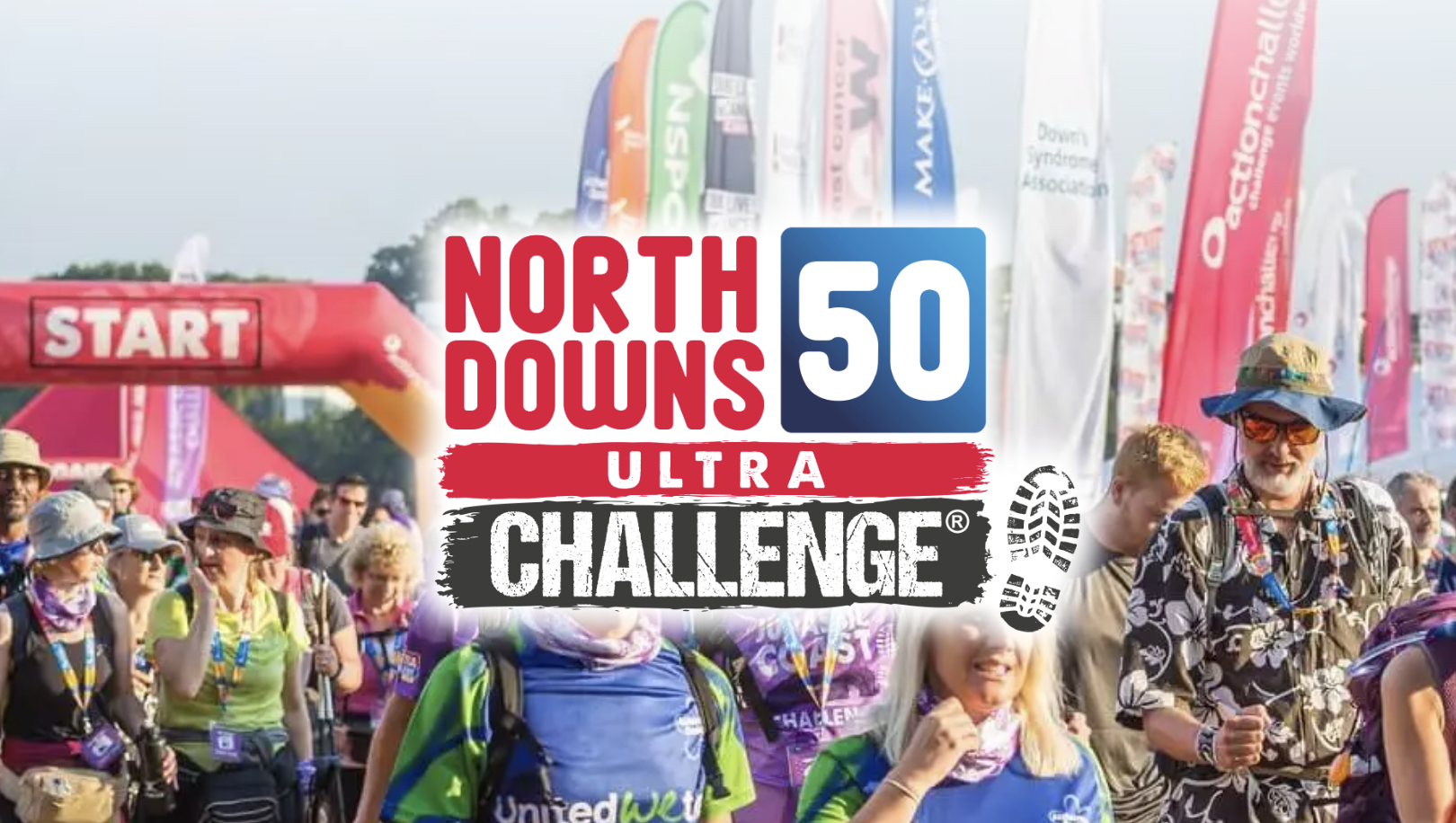 Excited participants gather at the starting line of the North Downs 50 Ultra Challenge, marked by a vibrant red 'Start' arch and colourful charity flags in the background. The crowd is filled with walkers and runners ready to take on the challenge, dressed in event gear and hats under sunny skies.