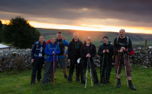 A group of six hikers standing together outdoors at sunset, smiling for the camera. They are equipped with hiking poles and dressed in outdoor gear. The backdrop features a scenic countryside landscape with rolling hills and a dramatic cloudy sky illuminated by the setting sun.