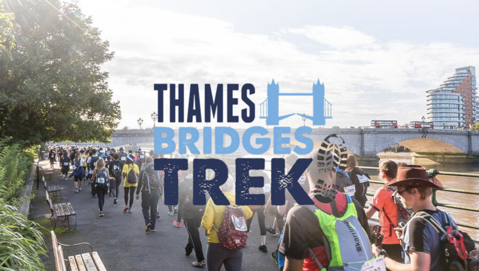 A group of people participating in the Thames Bridges Trek, walking along a riverside path under sunny skies. The event logo, featuring the words "Thames Bridges Trek" with an illustration of a bridge and a boot print, is overlaid on the image. A bridge and modern buildings can be seen in the background.